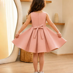 Young girl wearing a pink dress with a bow, standing in a room with wooden flooring and a shelf in the background.