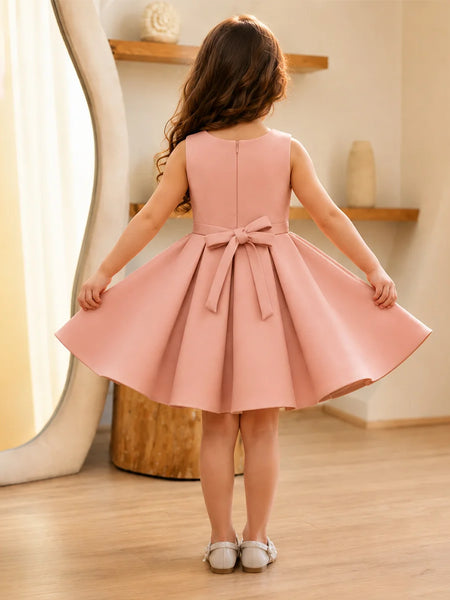 Young girl wearing a pink dress with a bow, standing in a room with wooden flooring and a shelf in the background.