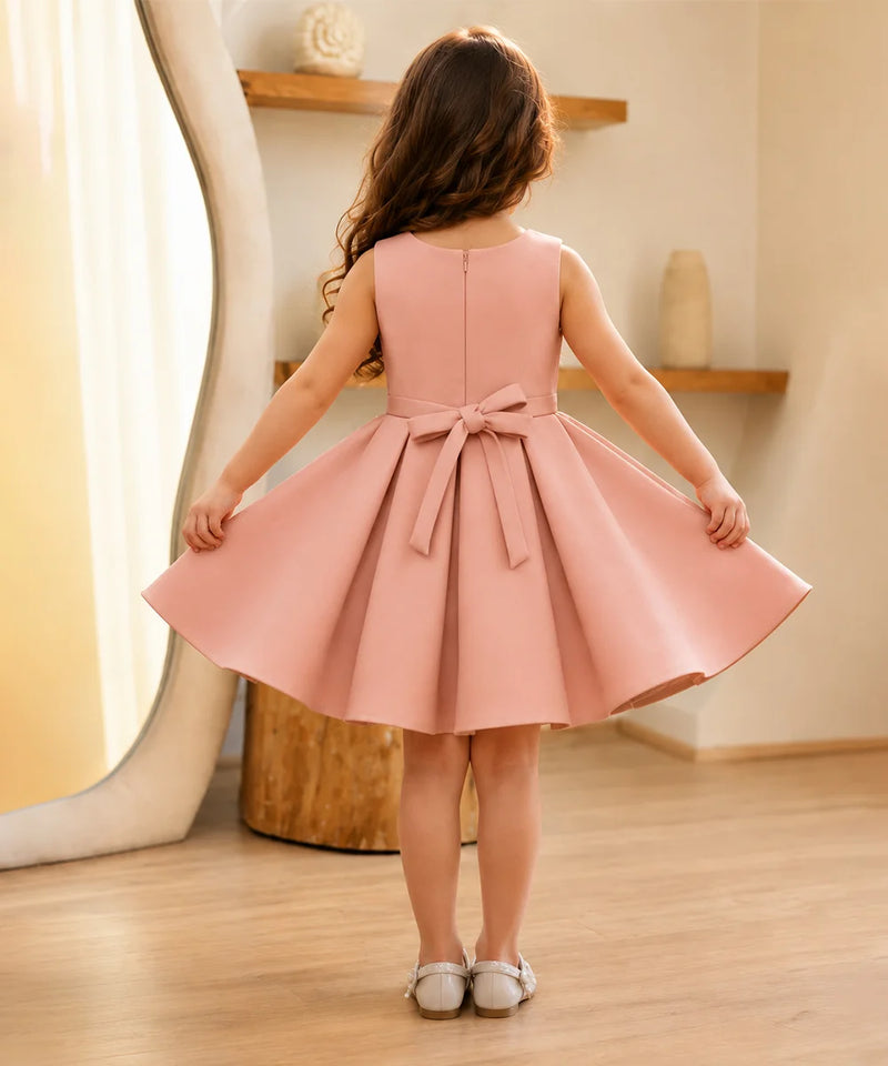 Young girl wearing a pink dress with a bow, standing in a room with wooden flooring and a shelf in the background.