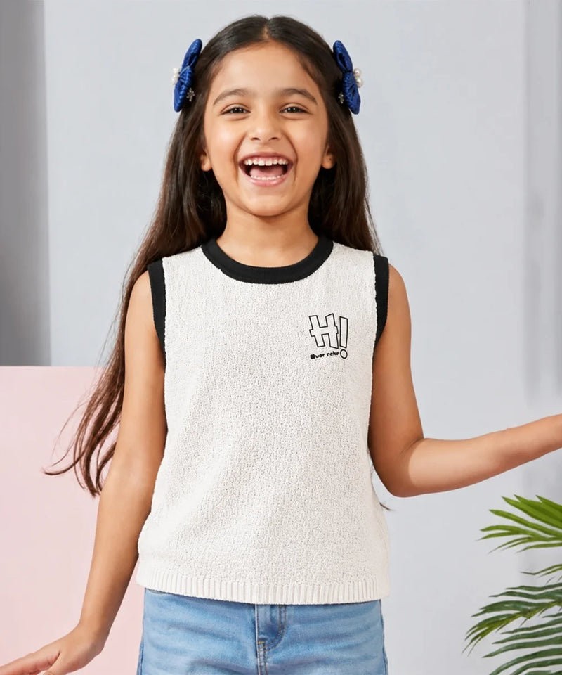 Young girl wearing a white sleeveless sweater with black trim and blue jeans, standing against a light gray background.