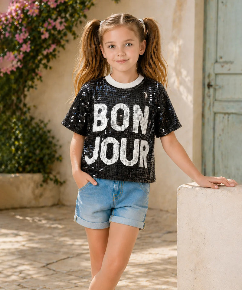 Young girl wearing a black sequin 'BON JOUR' t-shirt in an outdoor setting.
