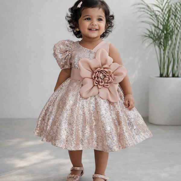 Young girl in a sparkly dress with a large pink flower standing indoors.