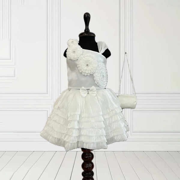 This White Coloured kids birthday wear dress consists of a crop top, a skirt and a matching handbag. It features floral detailing on the top and a cute bow on the waist.