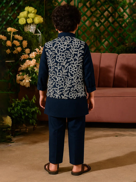 Person wearing a dark blue embroidered outfit standing in front of a pink sofa and decorative plants.