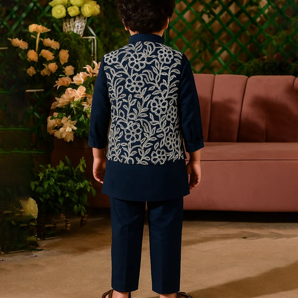 Person wearing a dark blue embroidered outfit standing in front of a pink sofa and decorative plants.