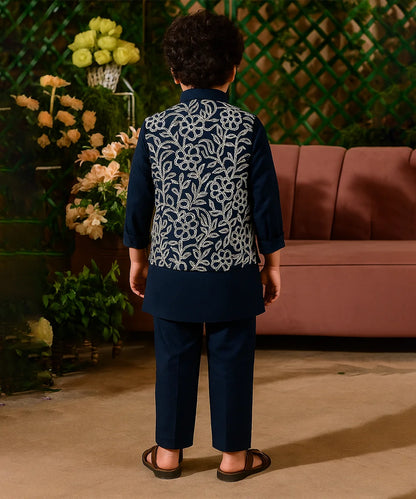 Person wearing a dark blue embroidered outfit standing in front of a pink sofa and decorative plants.