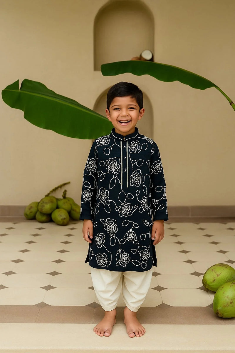 Child wearing a black kurta with white patterns standing in front of green leaves.