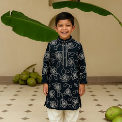 Child wearing a black kurta with white patterns standing in front of green leaves.