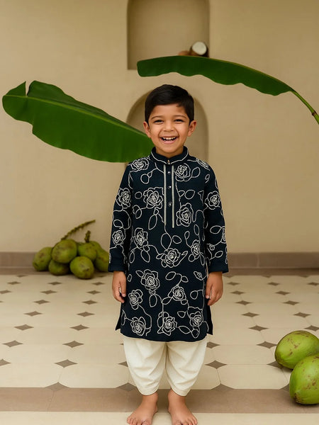 Child wearing a black kurta with white patterns standing in front of green leaves.