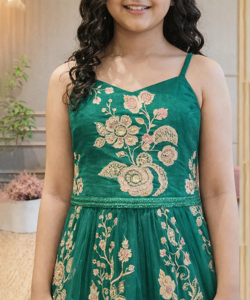 Young girl wearing a green embroidered dress with floral patterns indoors.