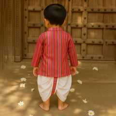 Child in a red and white striped traditional outfit standing in front of a wooden door with flowers on the ground.