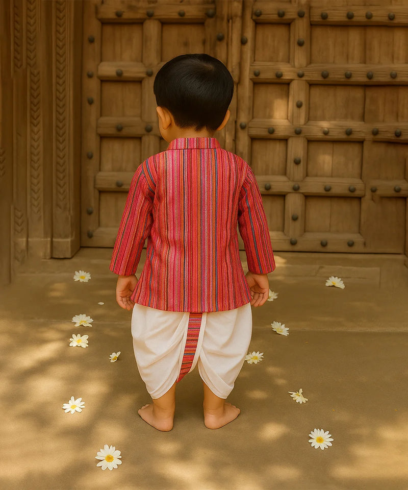 Child in a red and white striped traditional outfit standing in front of a wooden door with flowers on the ground.