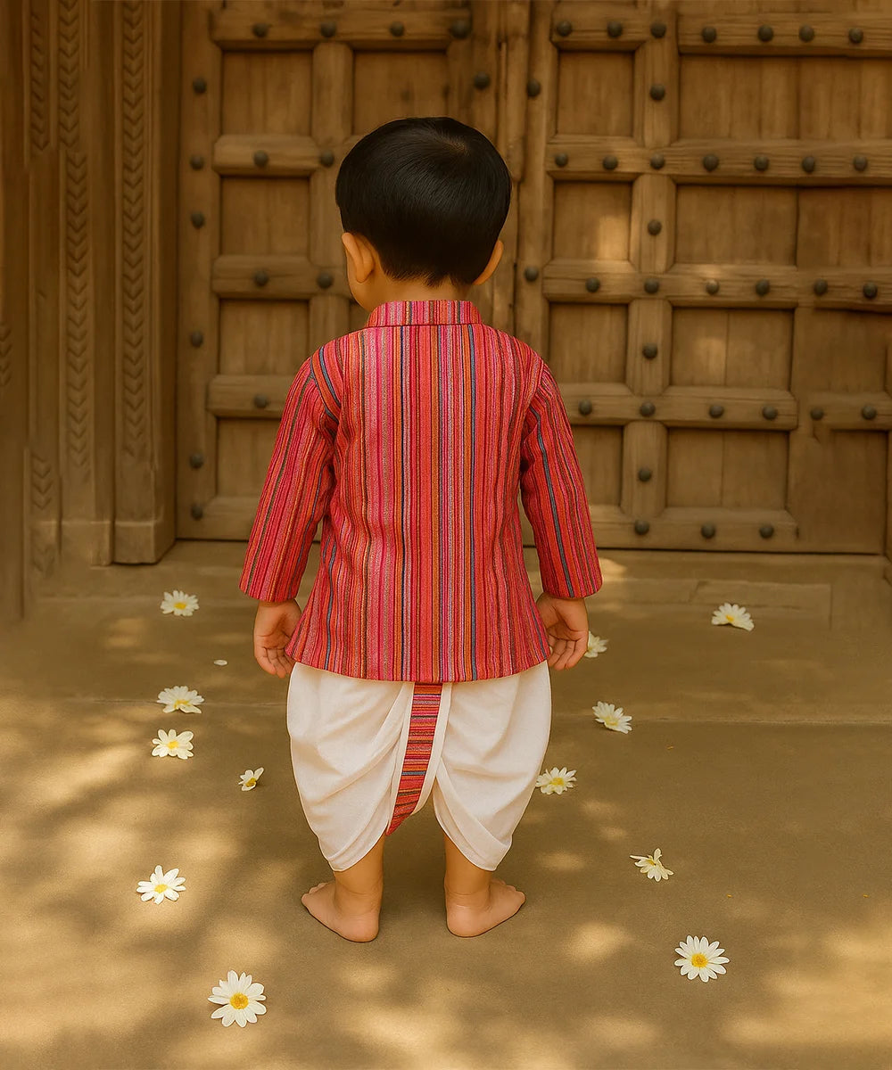 Child in a red and white striped traditional outfit standing in front of a wooden door with flowers on the ground.