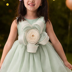 Young girl in a light green dress with floral details, standing outdoors with blurred lights in the background.