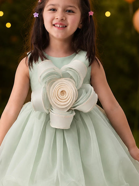 Young girl in a light green dress with floral details, standing outdoors with blurred lights in the background.