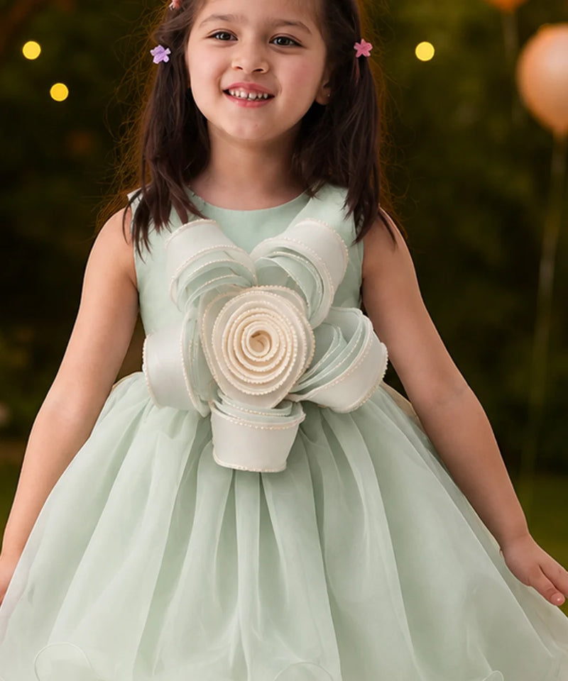 Young girl in a light green dress with floral details, standing outdoors with blurred lights in the background.