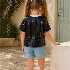 Young girl wearing a black sequin shirt and denim shorts standing outdoors.