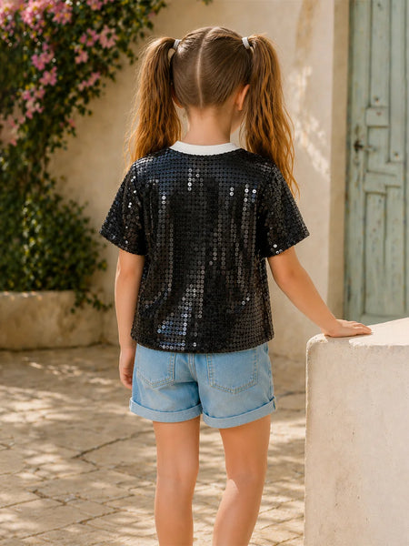 Young girl wearing a black sequin shirt and denim shorts standing outdoors.