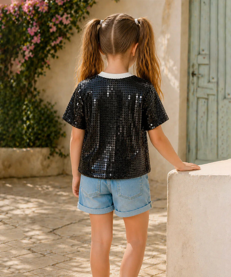 Young girl wearing a black sequin shirt and denim shorts standing outdoors.