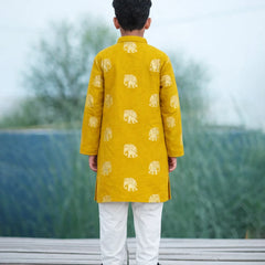 Child wearing a yellow kurta with elephant patterns and white pants, standing on a wooden platform with a blurred natural background.