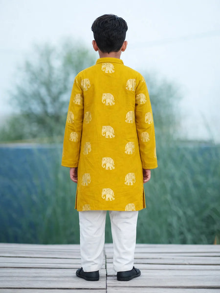 Child wearing a yellow kurta with elephant patterns and white pants, standing on a wooden platform with a blurred natural background.