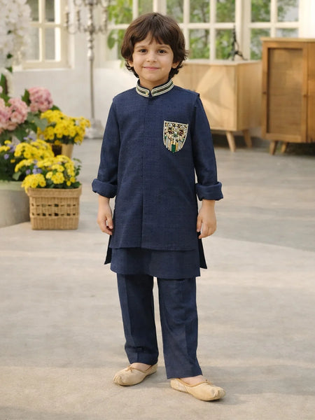Child wearing a navy blue traditional outfit with a decorative pocket, standing in a room with flowers and furniture.