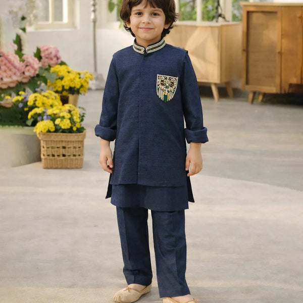 Child wearing a navy blue traditional outfit with a decorative pocket, standing in a room with flowers and furniture.