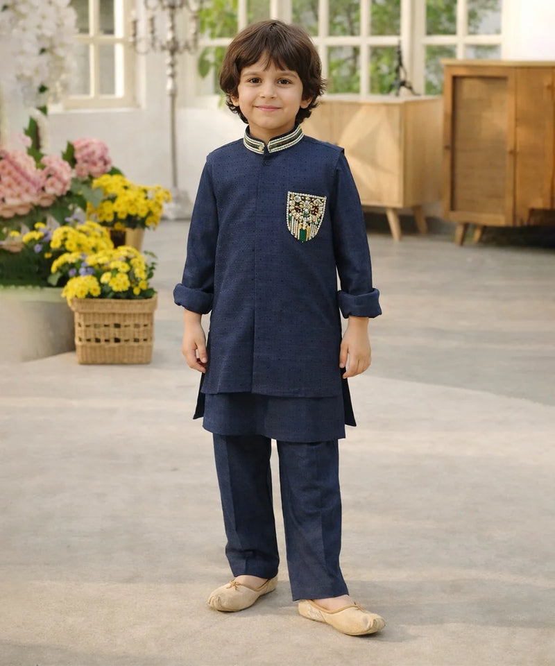 Child wearing a navy blue traditional outfit with a decorative pocket, standing in a room with flowers and furniture.