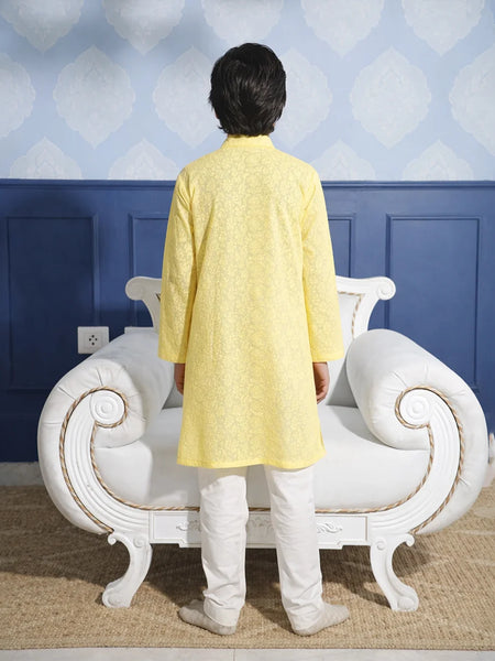Person wearing a yellow kurta with white pants standing in front of a decorative chair against a blue wall.