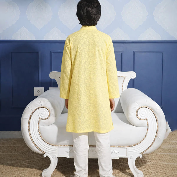 Person wearing a yellow kurta with white pants standing in front of a decorative chair against a blue wall.