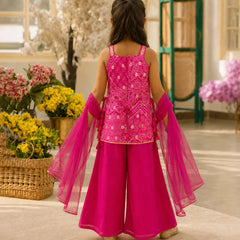 A young girl viewed from behind wearing a bright pink traditional outfit with a heavily embroidered sleeveless top, wide-leg pants, and sheer matching dupatta, standing in an indoor styled setting with flowers and green furniture.
