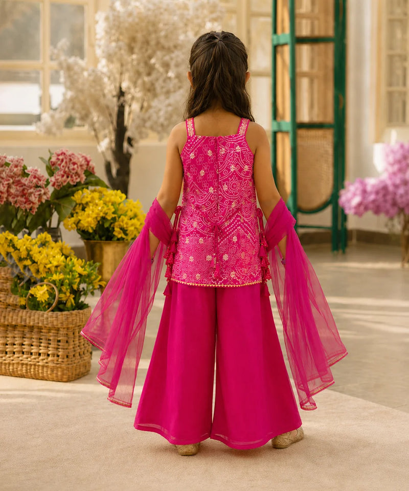 A young girl viewed from behind wearing a bright pink traditional outfit with a heavily embroidered sleeveless top, wide-leg pants, and sheer matching dupatta, standing in an indoor styled setting with flowers and green furniture.