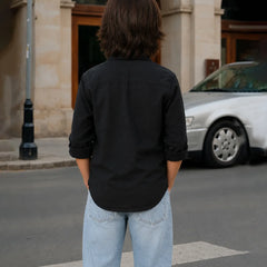Person wearing a black shirt and light blue jeans standing on a street with a car and building in the background.