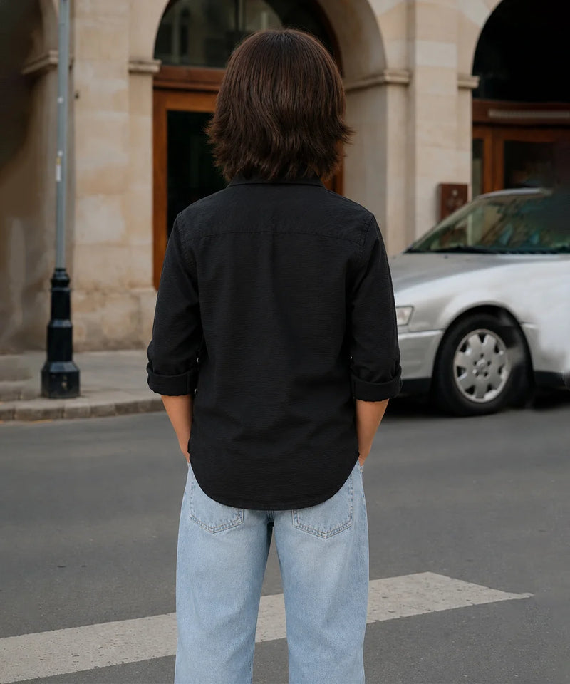 Person wearing a black shirt and light blue jeans standing on a street with a car and building in the background.