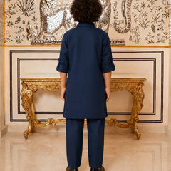 A person viewed from behind wearing a navy blue two-piece kurta and trouser set, standing in front of an ornate gold console table and a detailed wall tapestry.