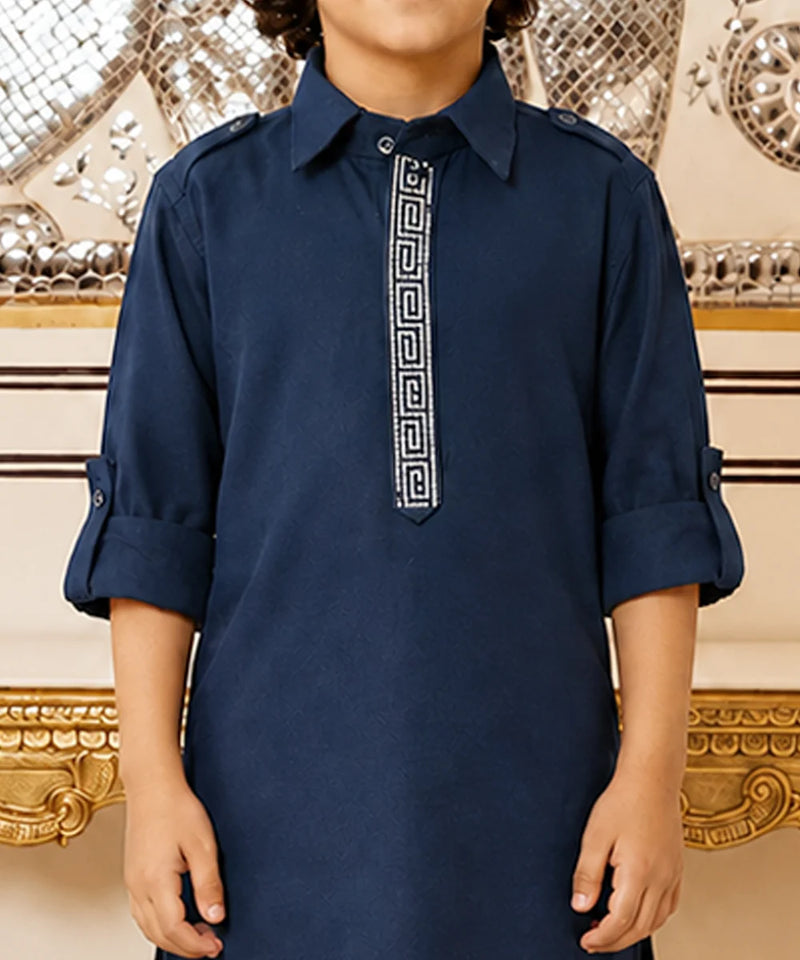 A child wearing a navy blue long-sleeve kurta with a white Greek key pattern down the center placket, standing in front of an ornate gold and silver mirror background.