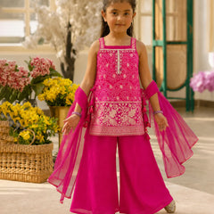 A young girl modeling a bright pink traditional three-piece outfit with intricate white embroidery, matching sheer pink arm sleeves, and wide-leg trousers, set against a warm, sunlit indoor background with flowers.