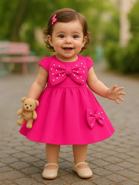 Child in a pink dress with a bow, holding a teddy bear, standing outdoors.