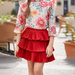 Young girl wearing a floral blouse and red skirt standing outdoors.