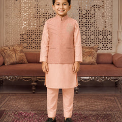 A young boy wearing a peach-colored traditional Indian outfit consisting of a sleeveless embroidered vest, kurta, and trousers, standing in a decorated room with a patterned screen and sofa in the background.