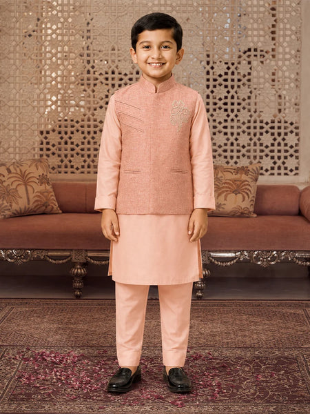 A young boy wearing a peach-colored traditional Indian outfit consisting of a sleeveless embroidered vest, kurta, and trousers, standing in a decorated room with a patterned screen and sofa in the background.