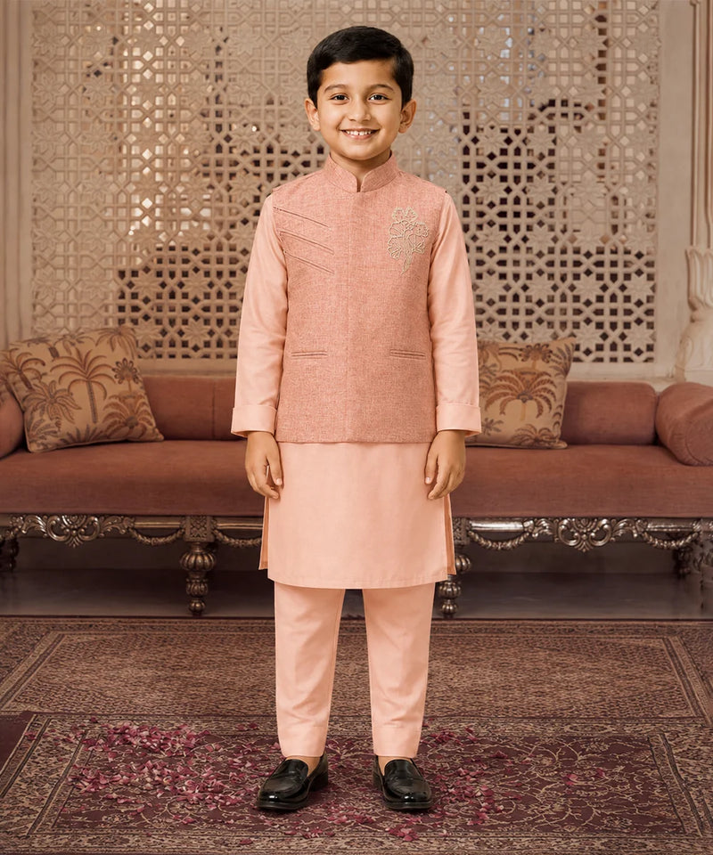 A young boy wearing a peach-colored traditional Indian outfit consisting of a sleeveless embroidered vest, kurta, and trousers, standing in a decorated room with a patterned screen and sofa in the background.
