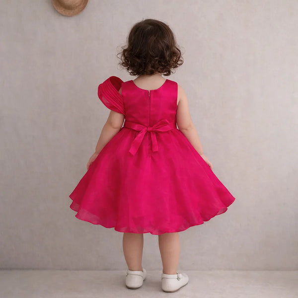 Child wearing a bright pink dress with a bow, standing against a plain background.