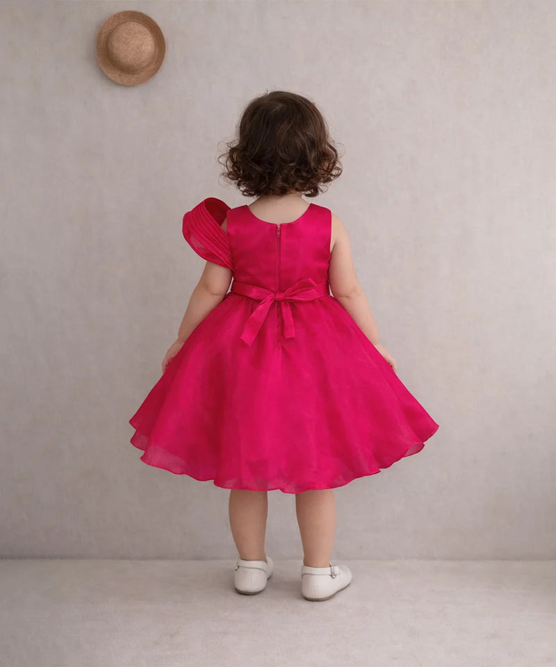 Child wearing a bright pink dress with a bow, standing against a plain background.