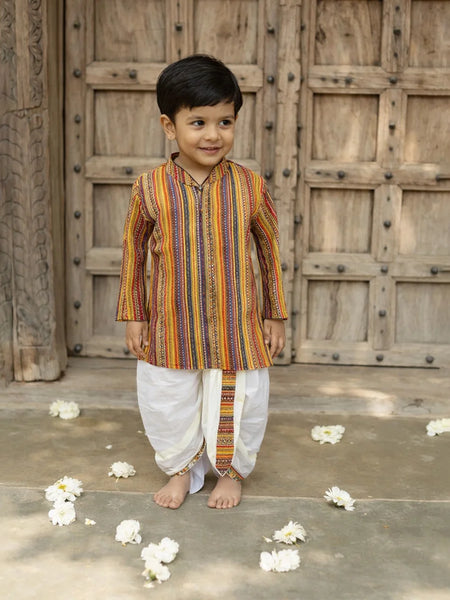 Young boy in a colorful kurta standing in front of a wooden door with flower petals on the ground.