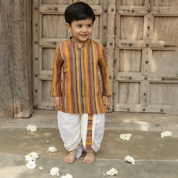 Young boy in a colorful kurta standing in front of a wooden door with flower petals on the ground.