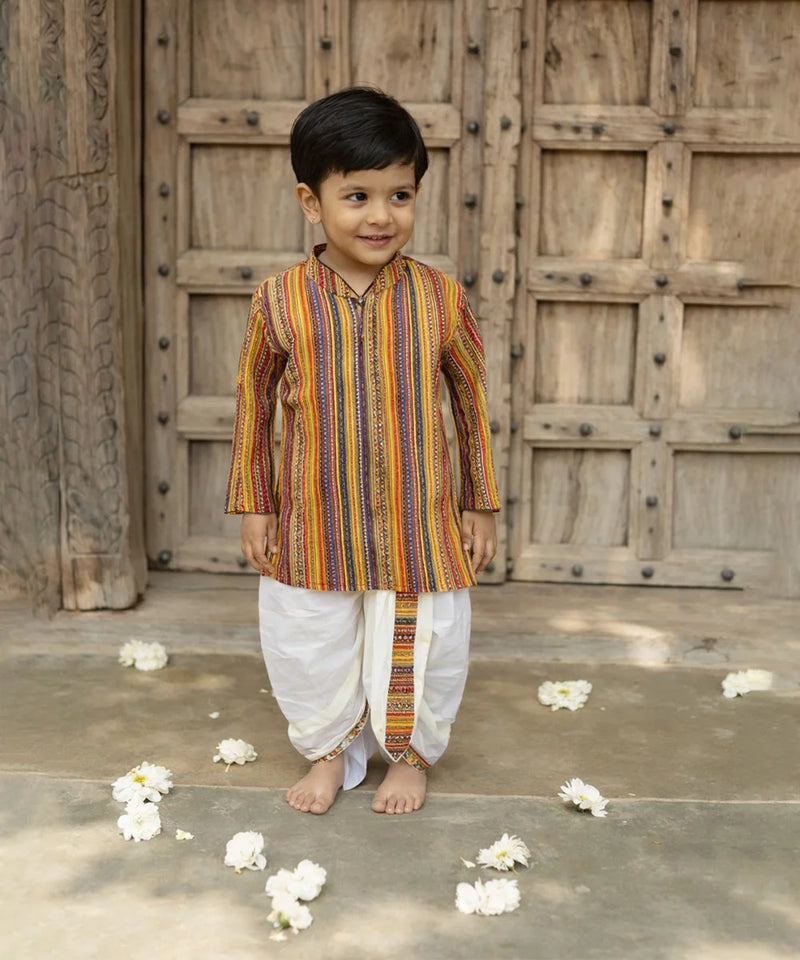 Young boy in a colorful kurta standing in front of a wooden door with flower petals on the ground.