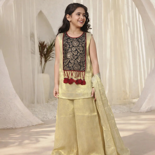 Young girl in a traditional outfit with a black embroidered top and light green palazzo pants, standing against a white curtain background.