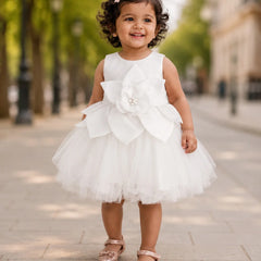 A toddler girl wearing a sleeveless white tulle dress with a large fabric flower applique on the front, standing on a paved outdoor walkway with blurred trees and buildings in the background.