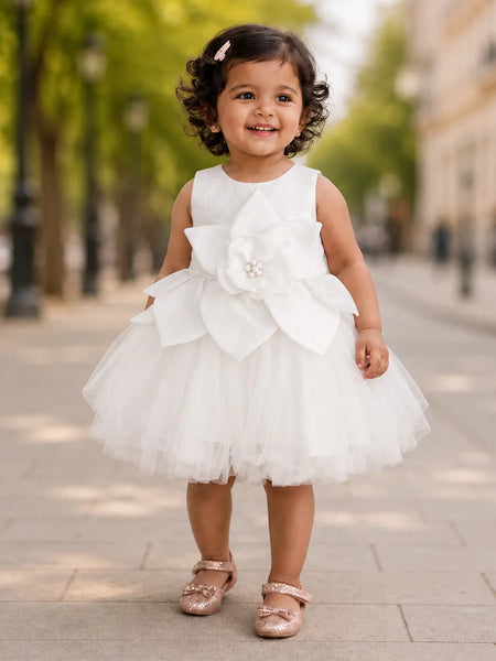 A toddler girl wearing a sleeveless white tulle dress with a large fabric flower applique on the front, standing on a paved outdoor walkway with blurred trees and buildings in the background.